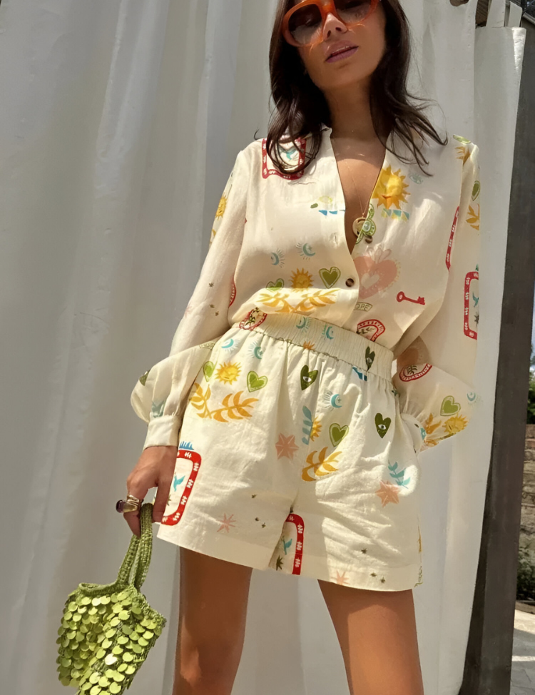 Donna con occhiali da sole indossa tuta estiva stampata con motivi floreali e geometrici, borsa verde a mano, sfondo bianco. Moda estiva femminile.
