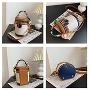 Borsa a tracolla cilindrica in pelle marrone e bianca con dettagli blu, manico colorato, su tavolo con lampada e giornale. Design elegante e moderno.
