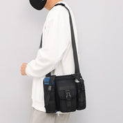 Uomo con felpa bianca indossa borsa a tracolla nera multifunzione, tasche multiple, stile casual urbano. Accessorio moda maschile pratico e trendy.