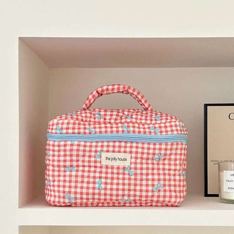 Borsa da toilette a quadretti rossi e bianchi con cerniera azzurra, manico superiore e motivo floreale, su mensola bianca.
