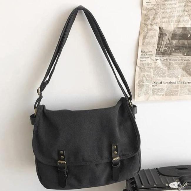 Borsa a tracolla nera in tela con fibbie, appesa a parete bianca. Design casual, ideale per uso quotidiano. Accessorio moda versatile.