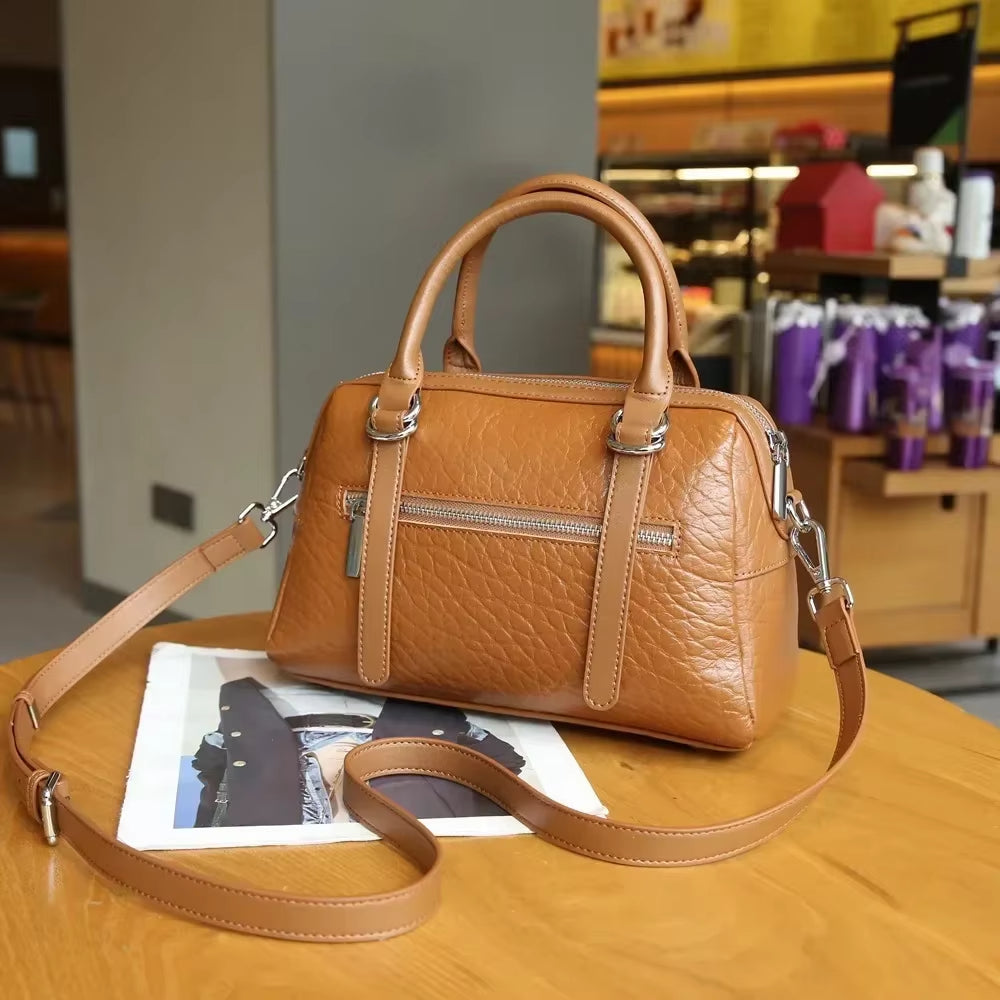 Borsa a mano in pelle marrone su tavolo di legno, con tracolla regolabile e tasca frontale con zip, in ambiente caffetteria. Accessorio moda elegante.