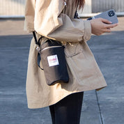 Giacca beige con borsa a tracolla nera, etichetta "Qidai", e smartphone in mano. Moda urbana, accessori pratici, stile casual.