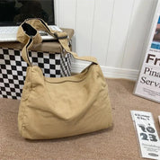 Borsa a tracolla beige in tessuto su pavimento marrone, accanto a scatola a scacchi e riviste. Design casual, ideale per uso quotidiano.
