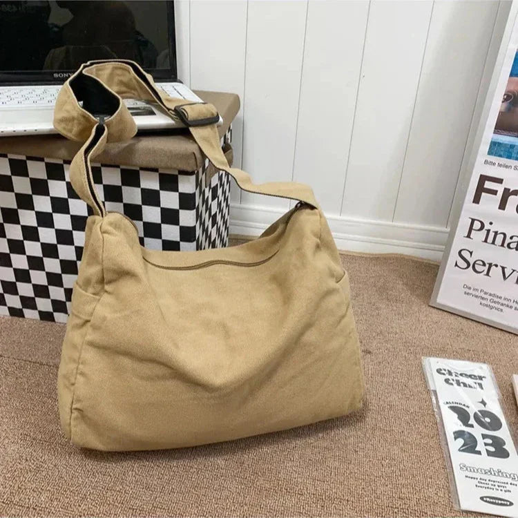 Borsa a tracolla beige in tessuto su pavimento marrone, accanto a scatola a scacchi e riviste. Design casual, ideale per uso quotidiano.