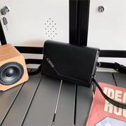 Borsa a tracolla nera elegante su tavolo grigio, accanto a speaker in legno e rivista. Accessorio moda donna, design moderno e versatile.