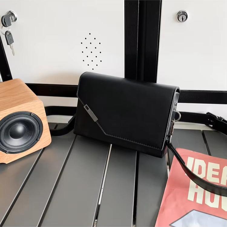 Borsa a tracolla nera elegante su tavolo grigio, accanto a speaker in legno e rivista. Accessorio moda donna, design moderno e versatile.