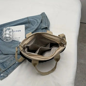 Borsa a tracolla beige aperta su letto bianco, accanto a giacca di jeans, libro e bicchiere d'acqua. Design elegante, moda donna, accessori casual.