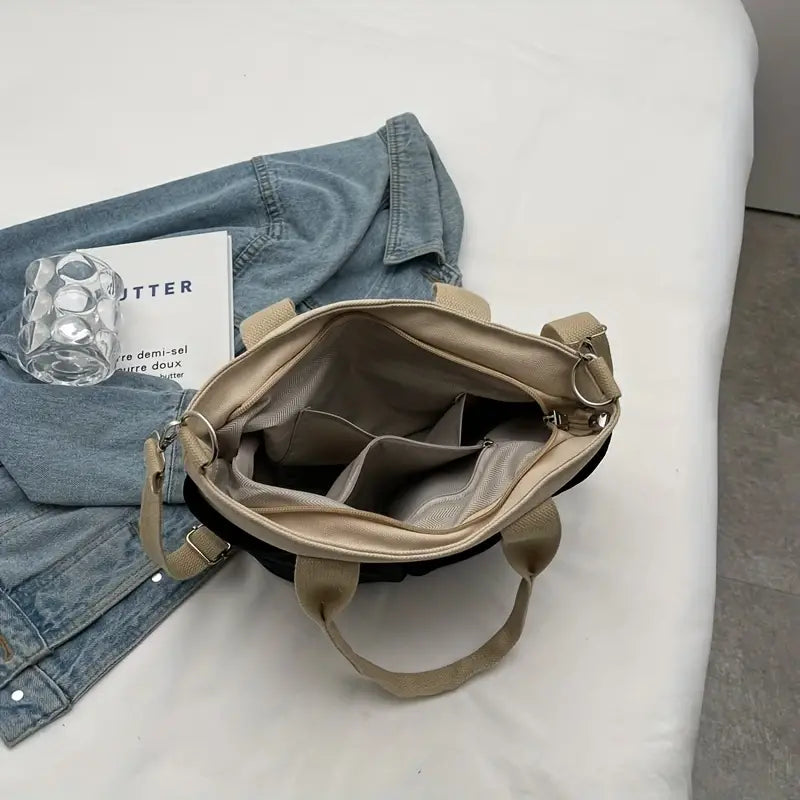 Borsa a tracolla beige aperta su letto bianco, accanto a giacca di jeans, libro e bicchiere d'acqua. Design elegante, moda donna, accessori casual.