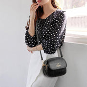 Donna con borsa a tracolla nera in pelle, indossa camicetta a pois e gonna bianca. Accessorio moda elegante, perfetto per look casual chic.