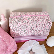 Beauty case rosa con motivo floreale, circondato da accessori da bagno: flacone spray, asciugamano rosa, fascia per capelli bianca. Organizzazione bagno elegante.