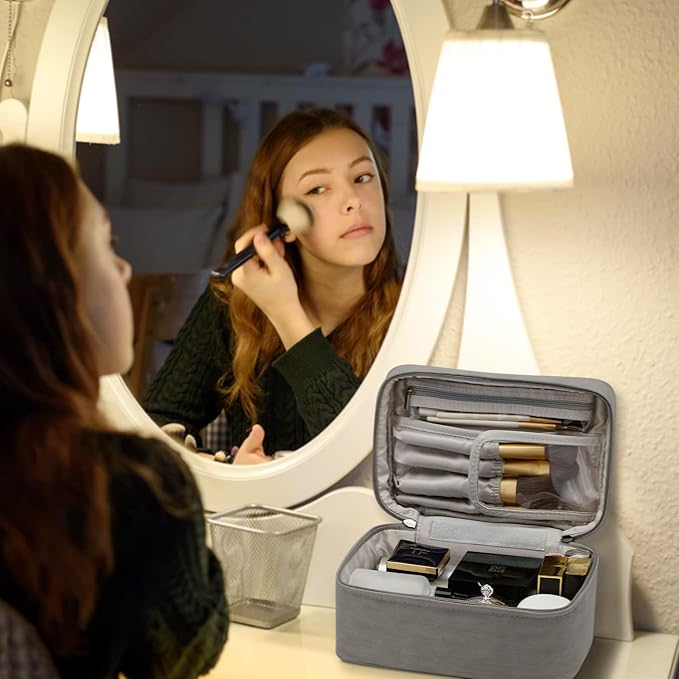 Donna si trucca davanti a uno specchio illuminato, con un beauty case grigio aperto contenente cosmetici. Accessori trucco, specchio, beauty case.