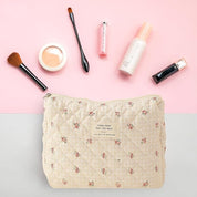 Beauty bag in tessuto trapuntato con motivo floreale su sfondo rosa, circondata da pennelli trucco, rossetto, fondotinta e crema viso.
