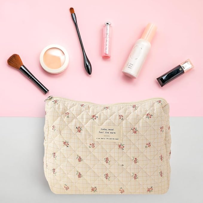 Beauty bag in tessuto trapuntato con motivo floreale su sfondo rosa, circondata da pennelli trucco, rossetto, fondotinta e crema viso.