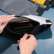 Mani che aprono una borsa a tracolla grigia, mostrando l'interno con portafoglio e accessori. Borsa resistente, ideale per viaggi e uso quotidiano.