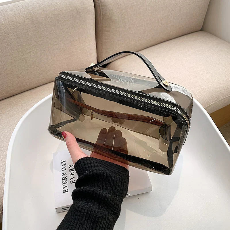 Borsa trasparente in PVC con cerniera, manico nero, su tavolo bianco. Design moderno, ideale per cosmetici e accessori. Elegante e funzionale.