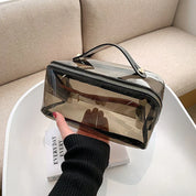Borsa trasparente in PVC con cerniera, manico nero, su tavolo bianco. Design moderno, ideale per cosmetici e accessori. Elegante e funzionale.