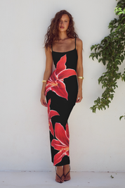 Modella con abito lungo nero a fiori rossi, spalline sottili, posa contro muro bianco con pianta verde. Moda estiva elegante, abbigliamento donna.