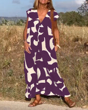 Donna con abito lungo viola e bianco astratto, sandali marroni, in un ambiente naturale. Moda estiva, abbigliamento casual elegante.