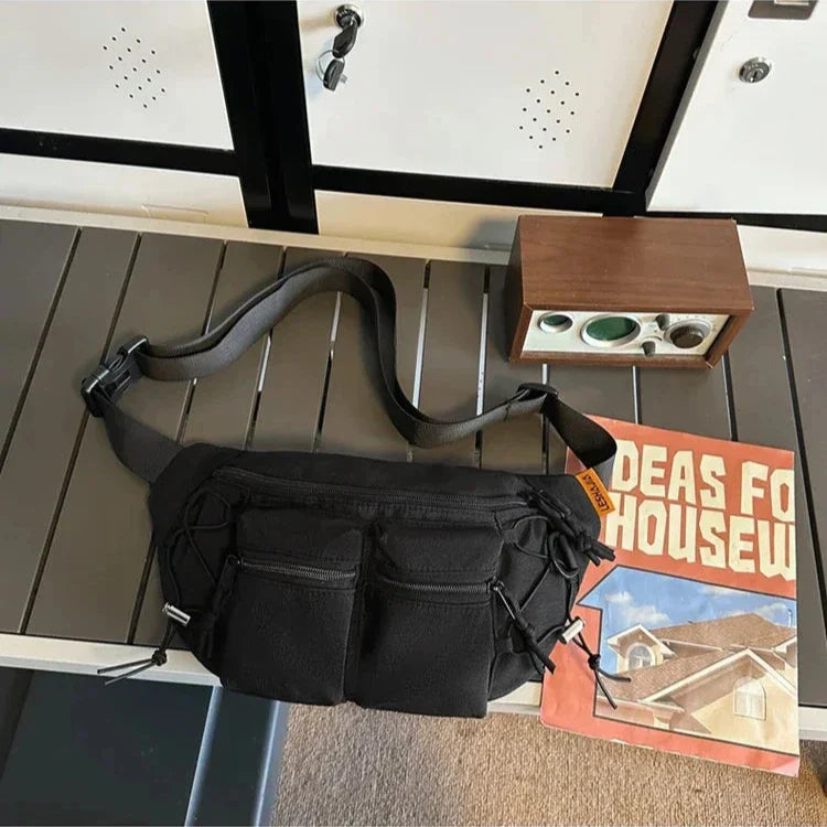 Marsupio nero con tasche multiple su tavolo grigio, accanto a rivista "Ideas for Housew" e radio vintage. Accessorio moda pratico e versatile.