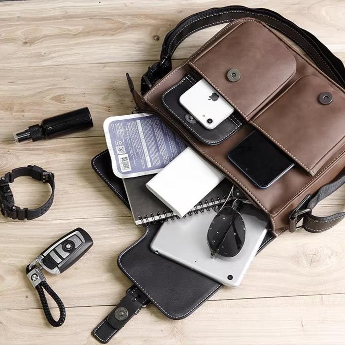 Borsa a tracolla in pelle marrone su tavolo di legno, contenente smartphone, occhiali da sole, taccuino, tablet e accessori. Elegante e funzionale.