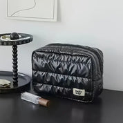 Borsa da toilette nera trapuntata su tavolo moderno, con etichetta "hello trip", accanto a vassoio con perle e profumo. Design elegante e funzionale.