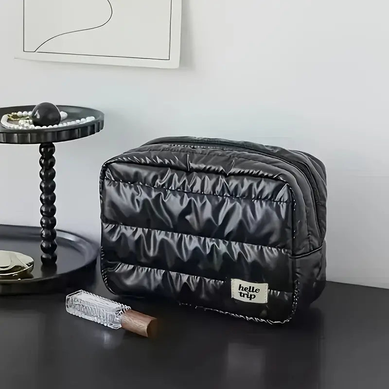 Borsa da toilette nera trapuntata su tavolo moderno, con etichetta "hello trip", accanto a vassoio con perle e profumo. Design elegante e funzionale.