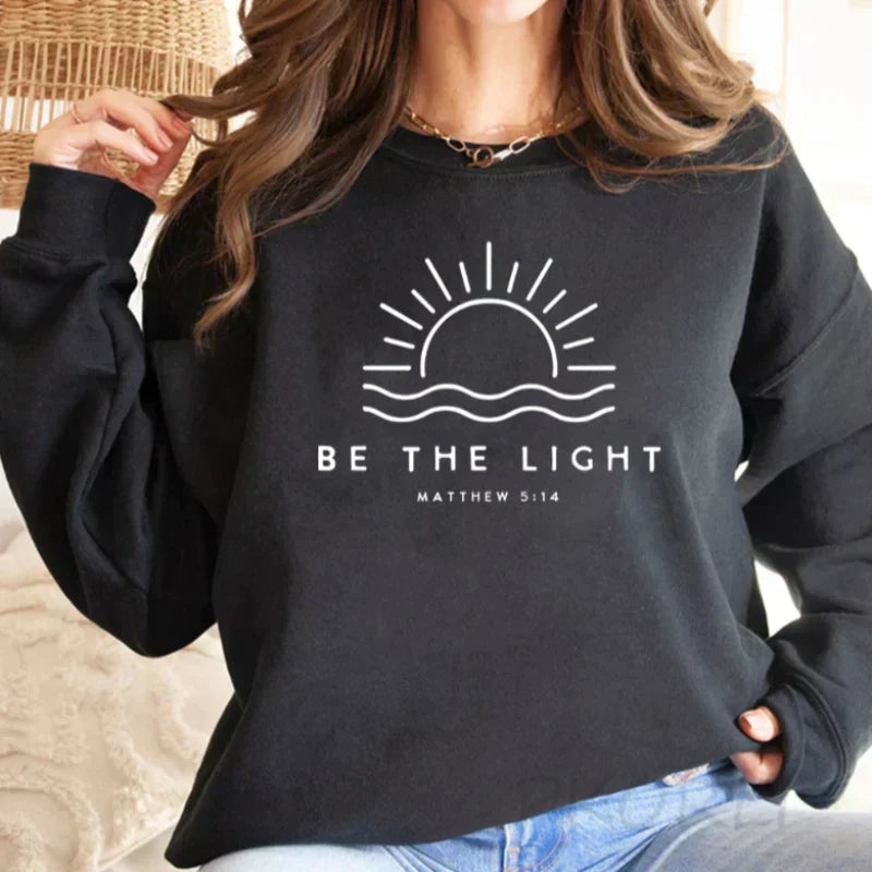 Felpa nera con grafica sole e scritta "Be the Light" e "Matthew 5:14", moda casual, abbigliamento ispirato alla fede, donna, stile confortevole.