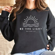 Felpa nera con grafica sole e scritta "Be the Light" e "Matthew 5:14", moda casual, abbigliamento ispirato alla fede, donna, stile confortevole.