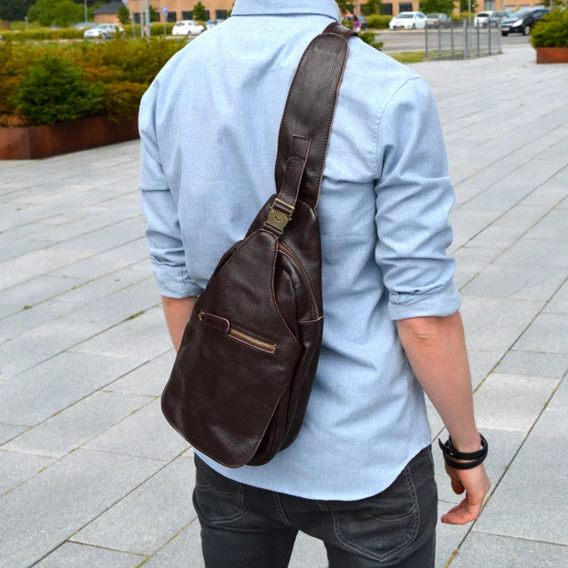 Uomo con zaino a tracolla in pelle marrone, camicia azzurra e jeans neri, all'aperto su pavimentazione grigia. Accessorio moda maschile elegante.