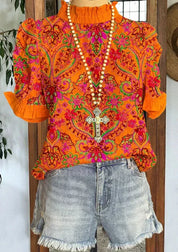 Blusa arancione a fiori con maniche corte e colletto arricciato, abbinata a shorts di jeans sfrangiati. Collana con croce decorativa.