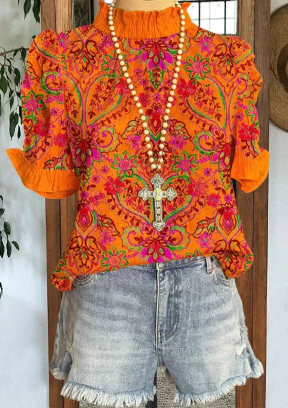 Blusa arancione a fiori con maniche corte e colletto arricciato, abbinata a shorts di jeans sfrangiati. Collana con croce decorativa.