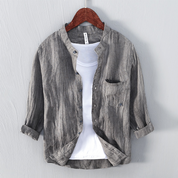 Camicia grigia in lino con colletto alla coreana, maniche lunghe arrotolate, abbinata a t-shirt bianca, appesa su gruccia di legno.