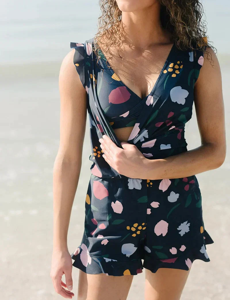 Donna in spiaggia con tuta corta floreale, scollo a V, colori vivaci. Moda estiva, abbigliamento casual, look trendy per il mare.