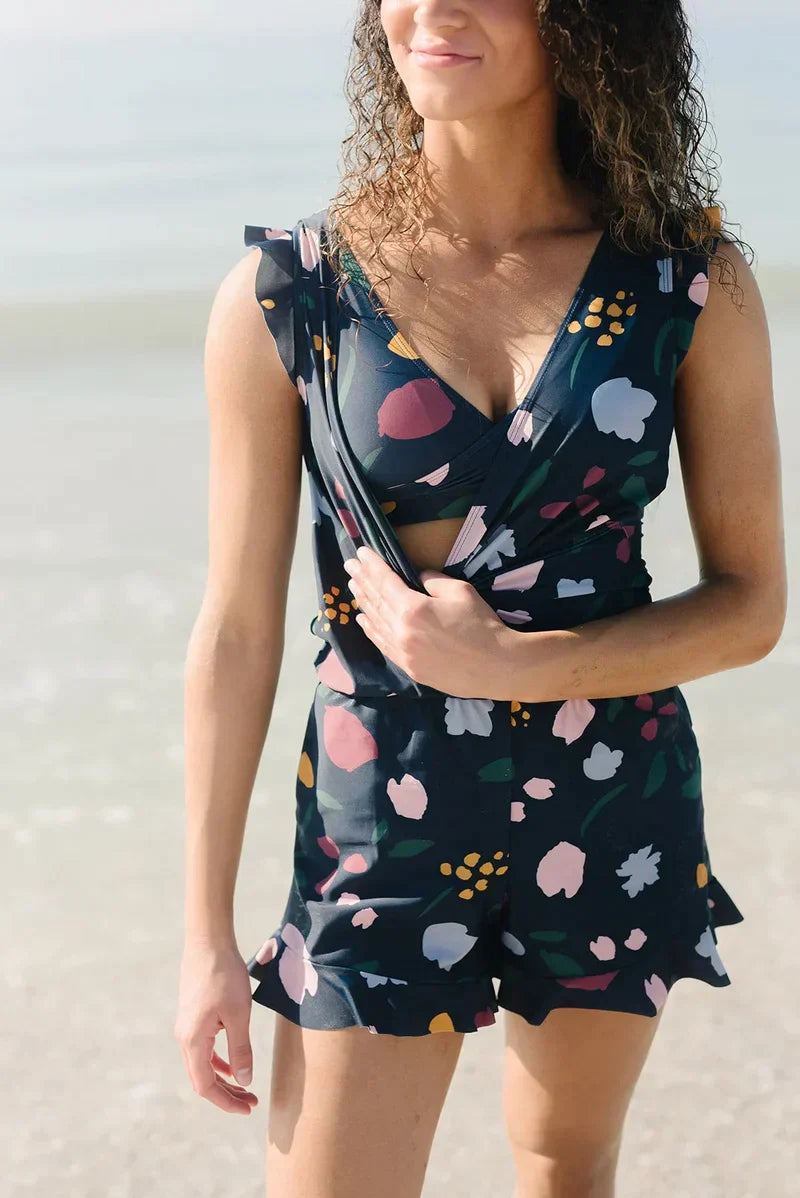 Donna in spiaggia con tuta corta floreale, scollo a V, colori vivaci. Moda estiva, abbigliamento casual, look trendy per il mare.
