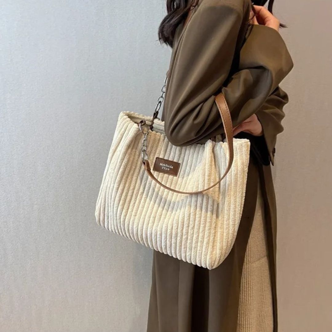Donna con cappotto marrone indossa borsa a spalla in tessuto beige a coste, manico in pelle marrone, elegante e moderna. Accessorio moda autunno.