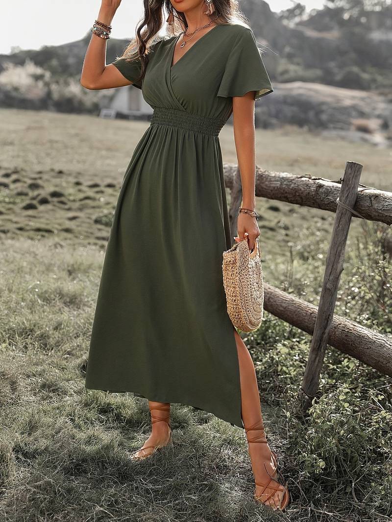 Donna con abito lungo verde oliva, cintura arricciata, maniche corte, sandali marroni, borsa di paglia, in un paesaggio rurale. Moda estiva elegante.