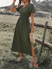 Donna con abito lungo verde oliva, cintura arricciata, maniche corte, sandali marroni, borsa di paglia, in un paesaggio rurale. Moda estiva elegante.