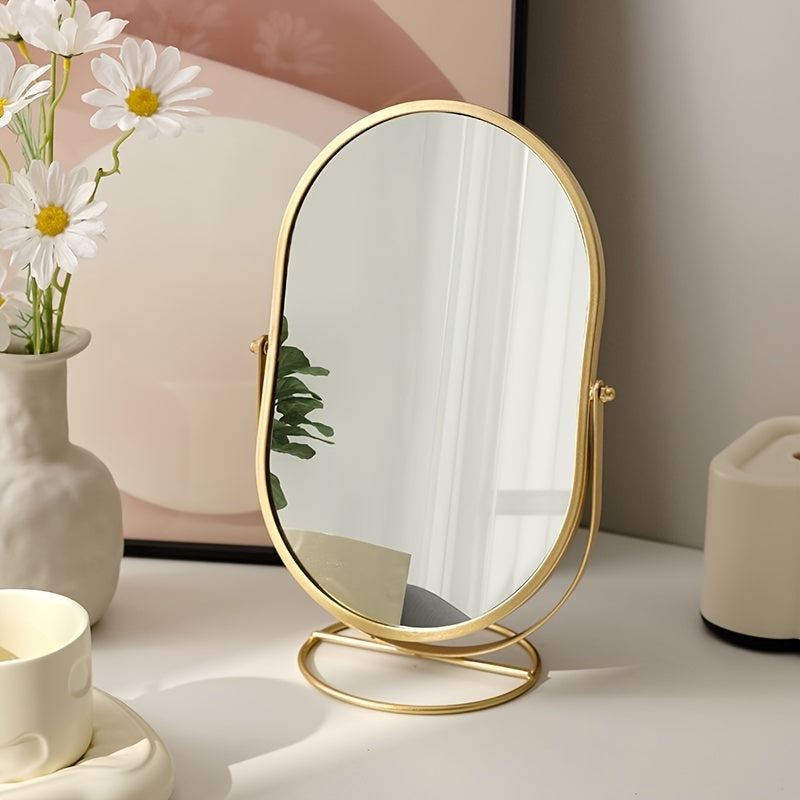 Specchio da tavolo ovale con cornice dorata, riflesso di piante verdi, accanto a vaso di fiori bianchi e tazza su scrivania moderna.