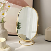 Specchio da tavolo ovale con cornice dorata, riflesso di piante verdi, accanto a vaso di fiori bianchi e tazza su scrivania moderna.