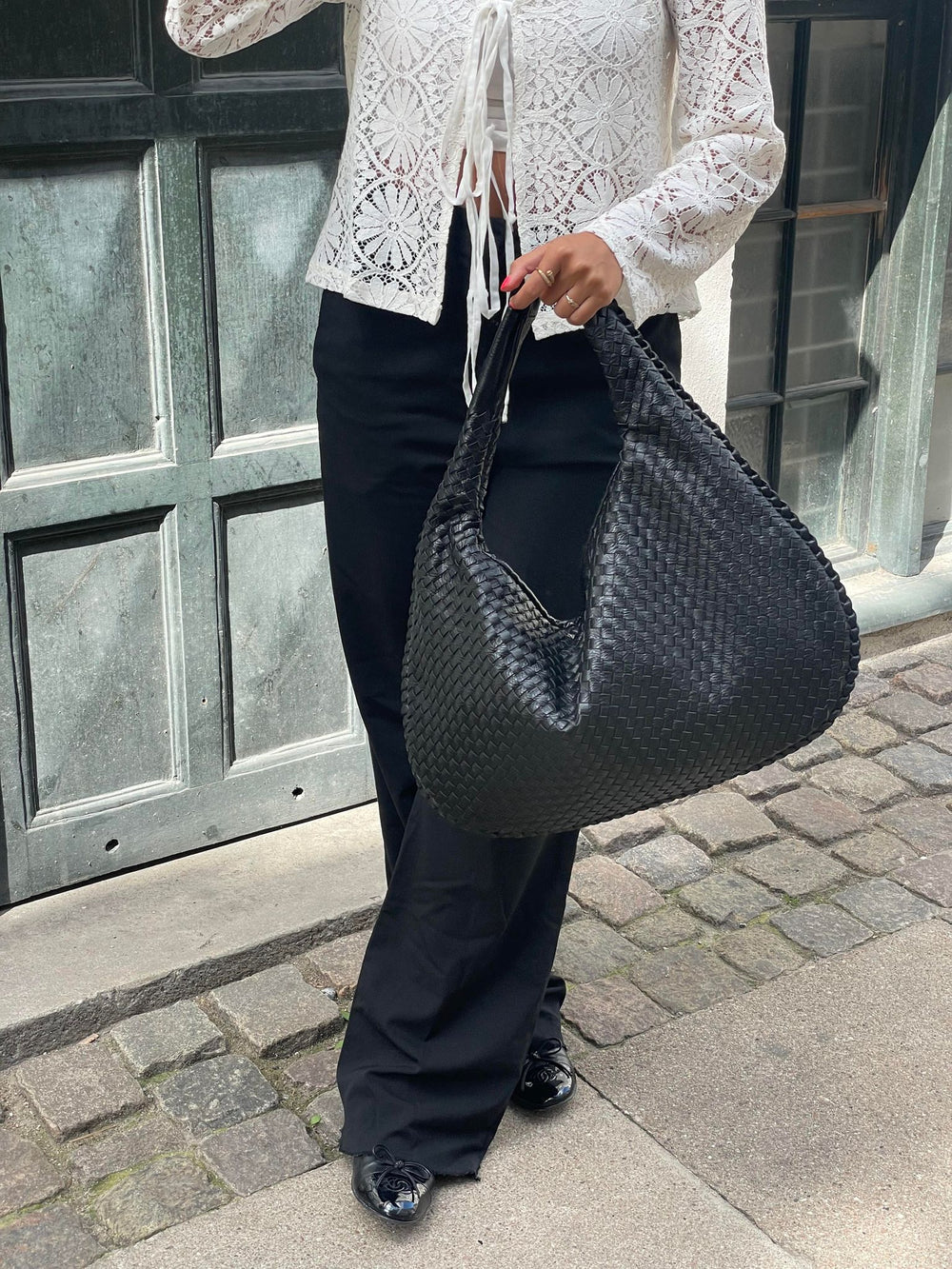 Donna con borsa a spalla intrecciata nera, indossa camicia bianca in pizzo e pantaloni neri. Moda urbana elegante, accessori di tendenza.