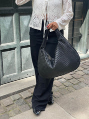 Donna con borsa a spalla intrecciata nera, indossa camicia bianca in pizzo e pantaloni neri. Moda urbana elegante, accessori di tendenza.