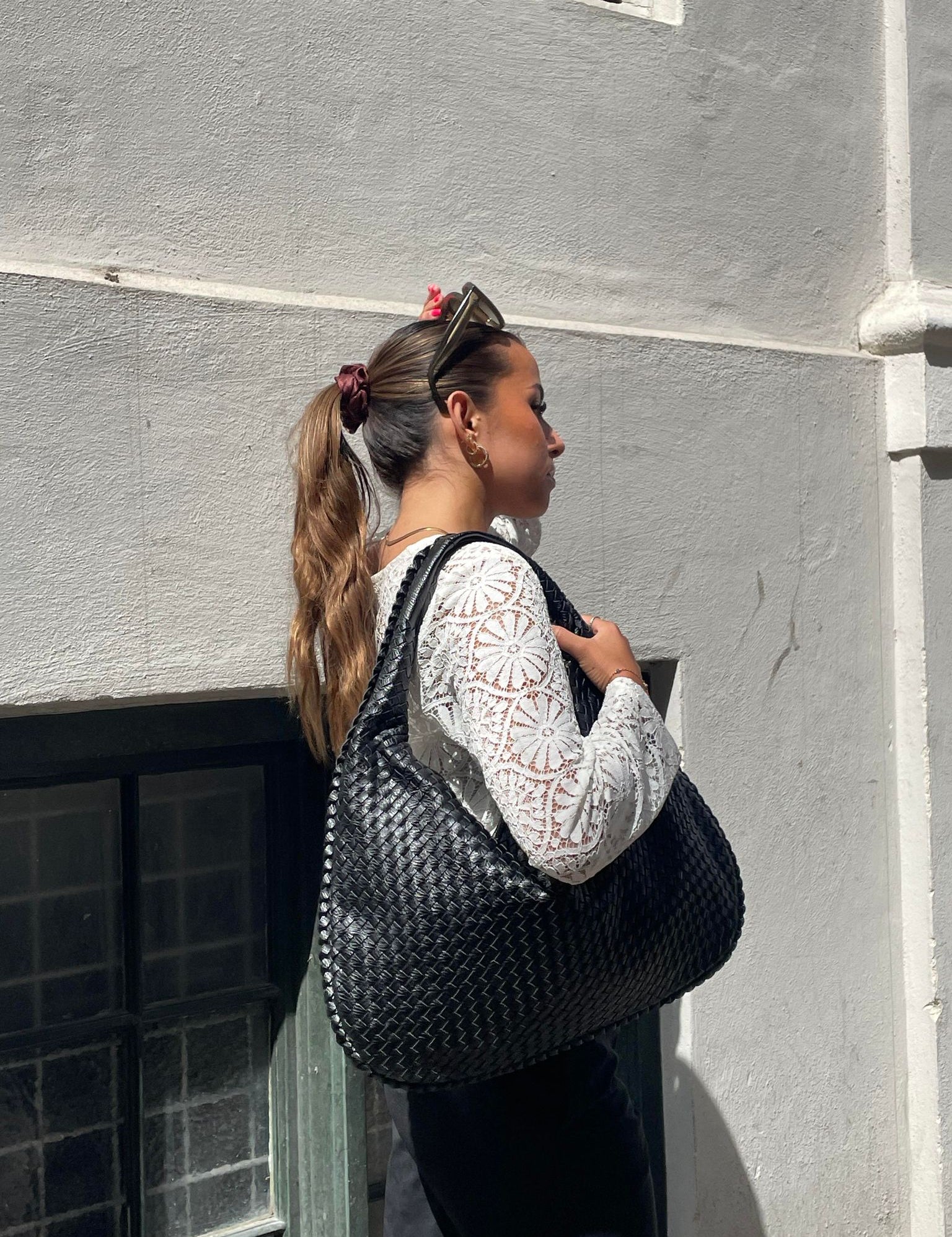 Donna con borsa intrecciata nera, indossa maglia bianca a maniche lunghe con motivo floreale. Moda urbana, accessori eleganti, stile casual chic.