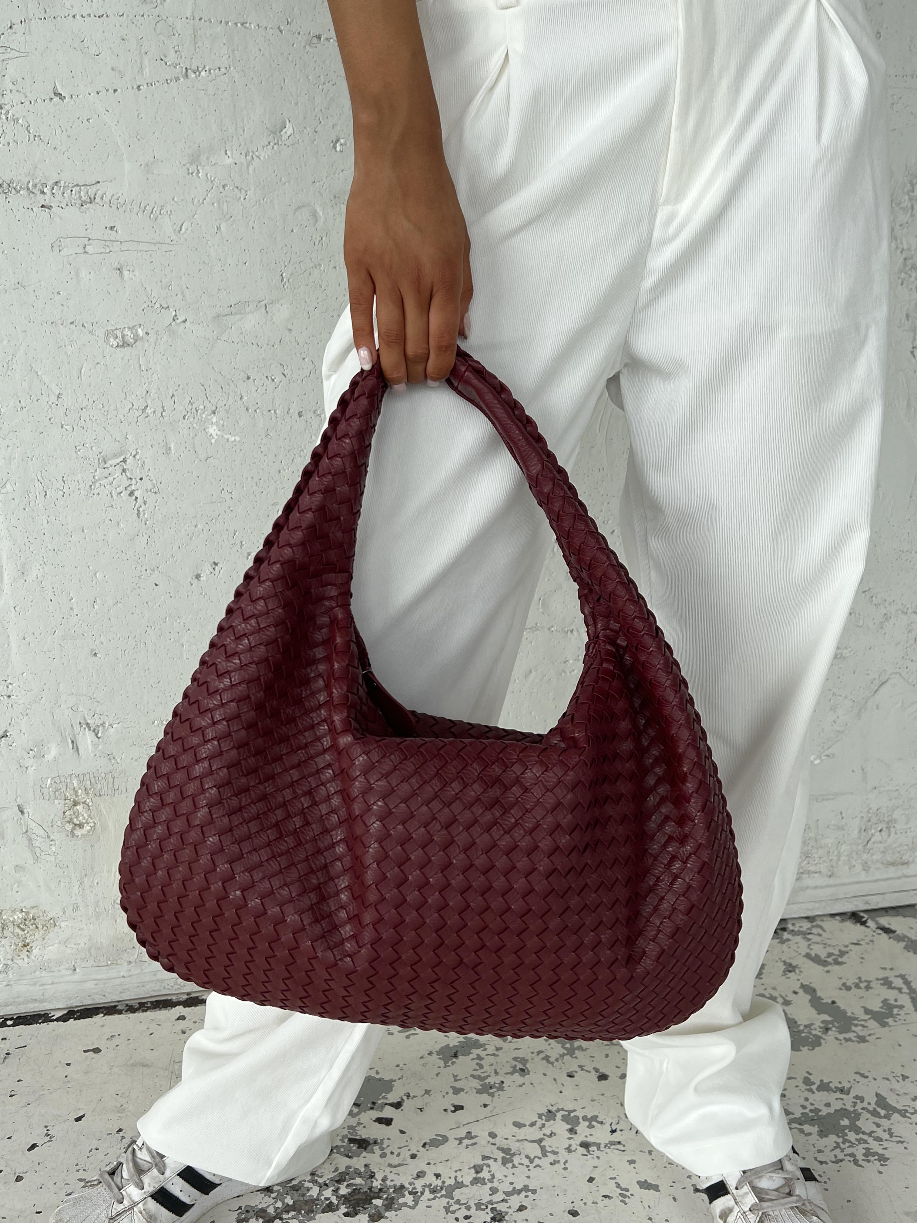 Borsa a spalla intrecciata in pelle bordeaux, indossata da una persona con pantaloni bianchi. Accessorio moda elegante e di tendenza.