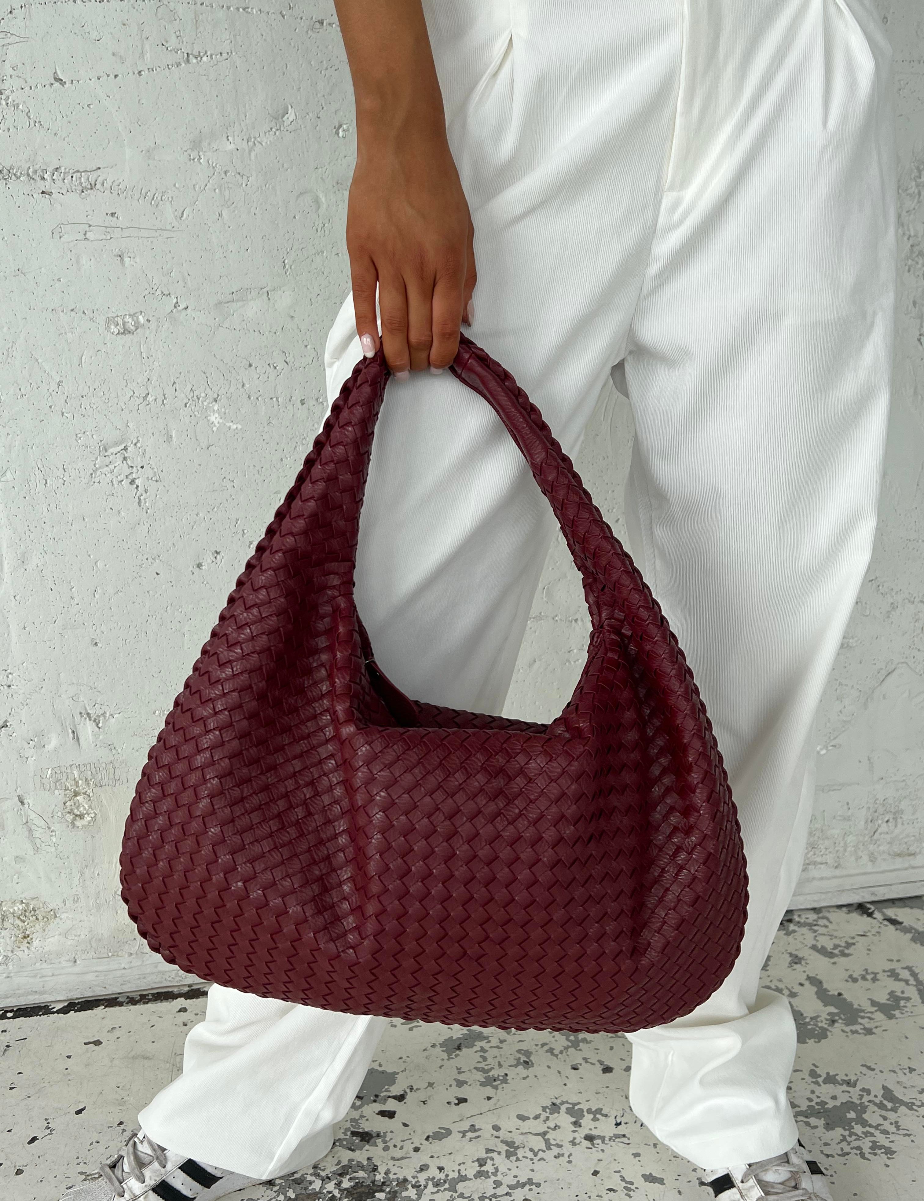 Borsa a spalla intrecciata in pelle bordeaux, indossata da una persona con pantaloni bianchi. Accessorio moda elegante e di tendenza.