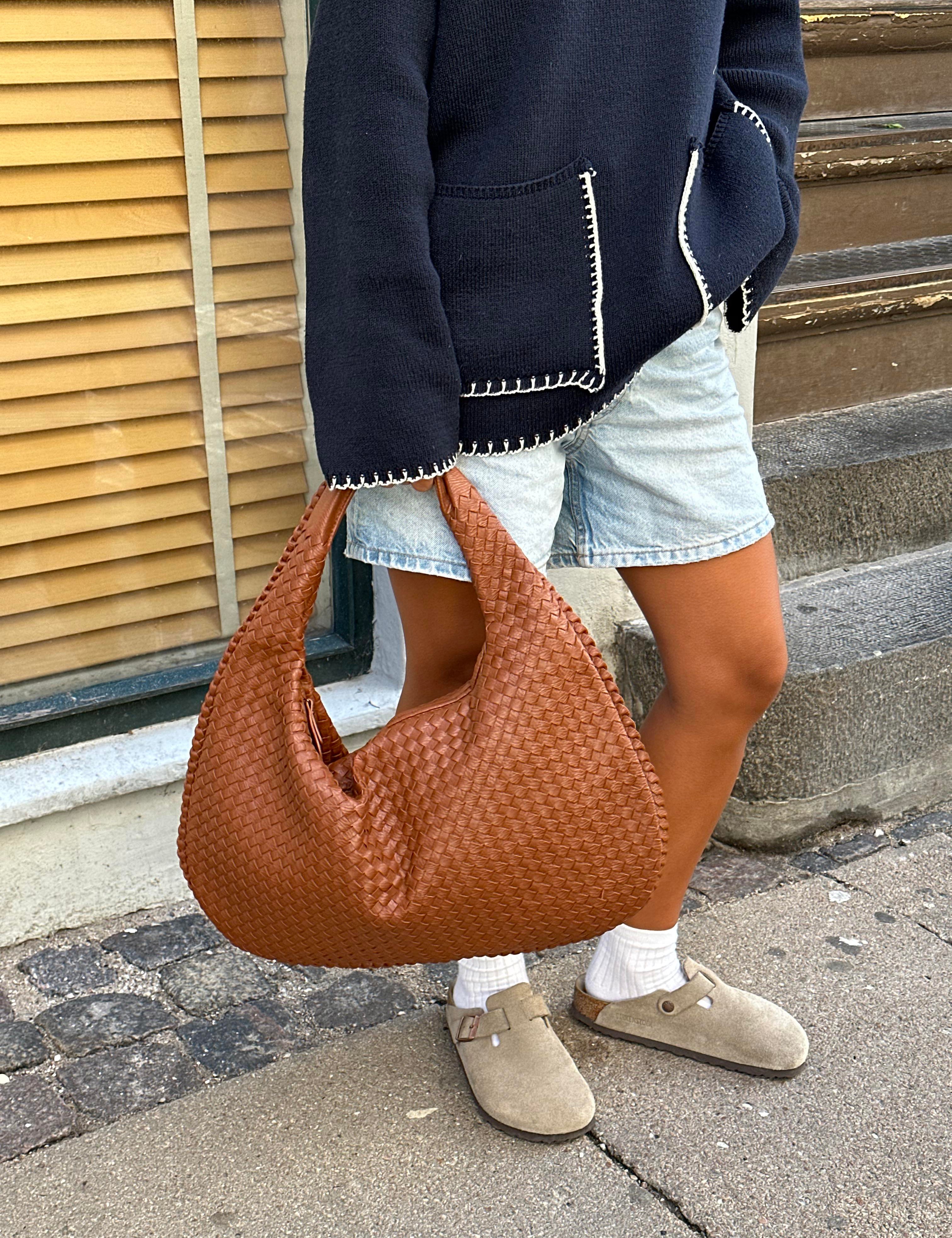Donna con borsa a spalla intrecciata marrone, indossa giacca blu e pantaloncini di jeans. Scarpe beige, look casual urbano.