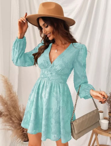 Donna con abito azzurro in pizzo, cappello beige, borsa a tracolla, sfondo con piante decorative. Moda femminile elegante e chic.