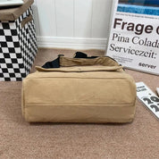 Borsa tote beige in tessuto su pavimento marrone, accanto a scatola a scacchi e poster. Design casual, ideale per shopping e uso quotidiano.
