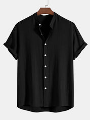 Camicia nera da uomo in cotone, maniche corte, colletto alla coreana, bottoni bianchi, stile casual elegante, appesa su gruccia di legno.
