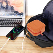 Custodia arancione per cavi su tavolo di legno, accanto a zaino nero, smartphone in carica e laptop. Accessorio organizzazione viaggio.